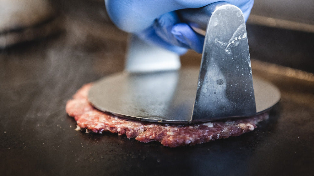 Hamburger patty being flattened with a meat tenderizer on a dark surface.